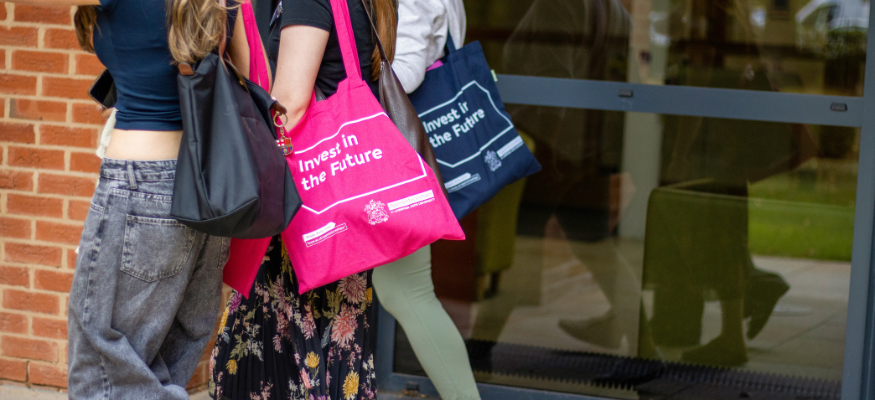 3 apprentices walking out of shot, with a pink and navy tote bag with the apprenticeships team slogan, Invest in the future, displayed to camera